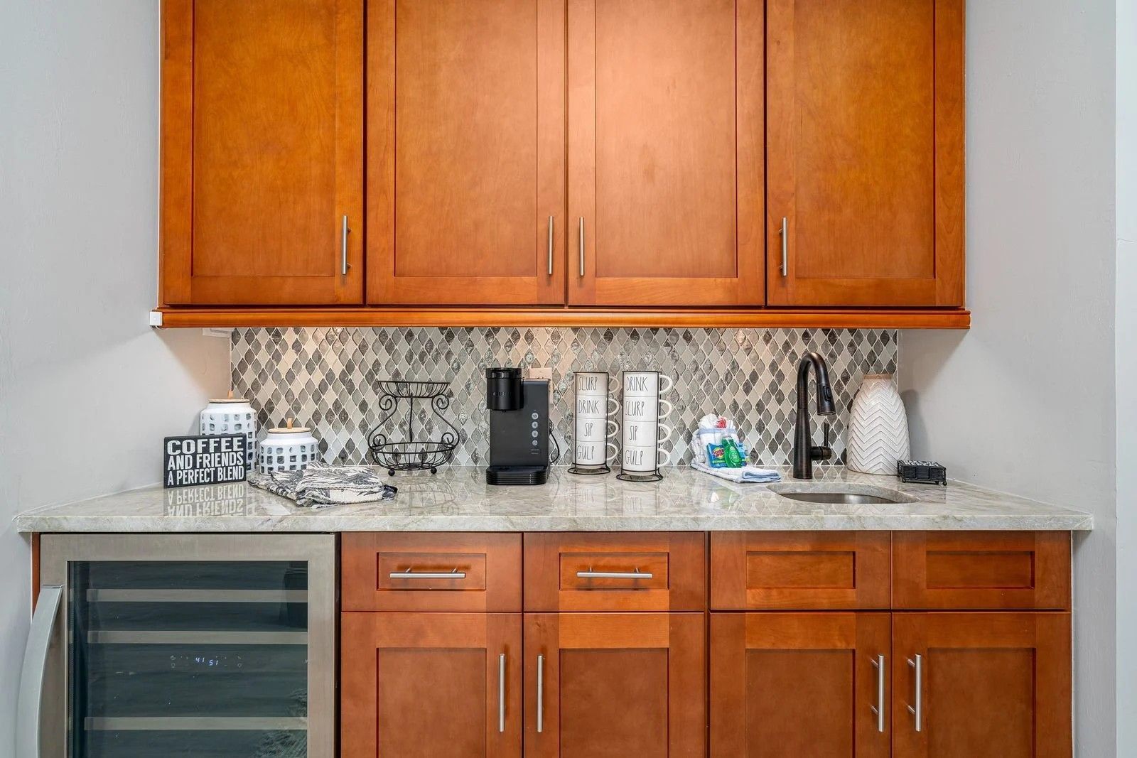 Modern kitchen counter with wooden cabinets, stone backsplash, coffee maker, and small wine fridge