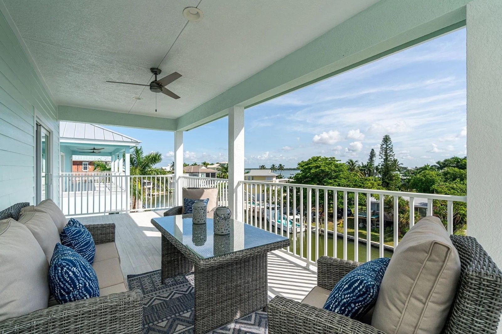 Covered balcony with cushioned seating, glass-top table, and white railing overlooking a green landscape.