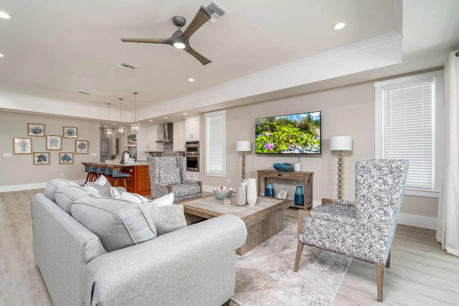 Bright open living room with gray sofa, patterned chairs, wall TV, ceiling fan, and kitchen in the background