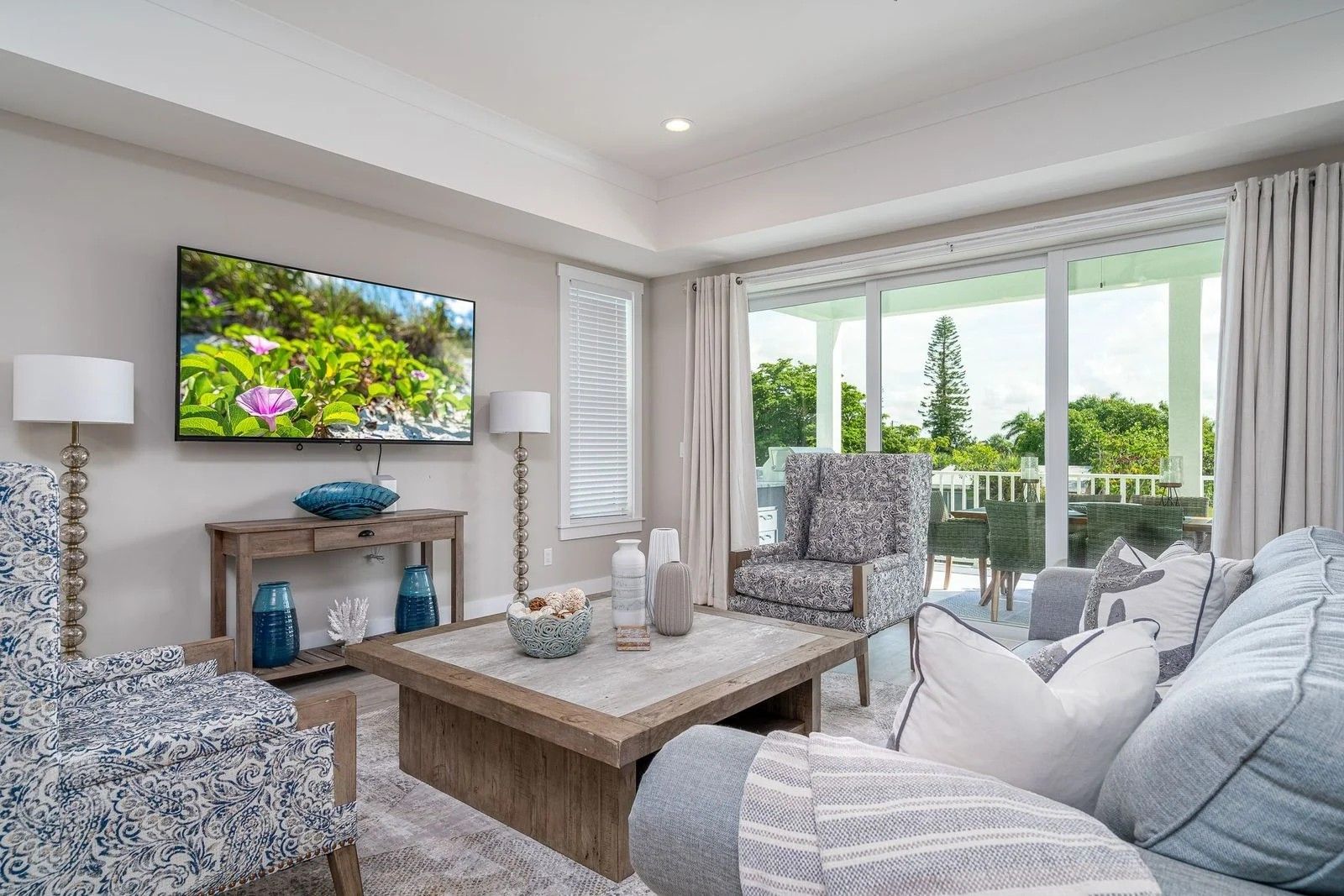 Bright living room with patterned chairs, a sofa, wall-mounted TV, and large windows overlooking greenery