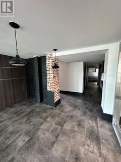 Interior view of a room with dark flooring, a brick accent column, and hanging black pendant lights. A hallway is visible in the background.
