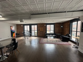 Spacious loft interior with exposed brick walls, large windows, and dark wood floors. Includes seating areas, a dining table, and decorative rug.