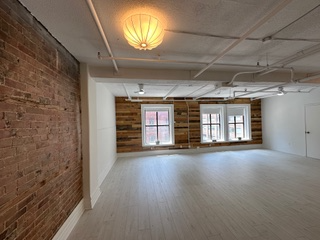 Empty, bright room with brick wall on left, white floor, two windows in the center, and a decorative light fixture.