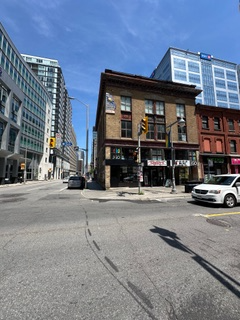 Street corner with a two-story brick building. Tall modern buildings are visible in the background under a blue sky.