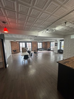 Spacious, open-concept loft with dark hardwood floors, exposed brick walls, and a decorative ceiling. Dining table with chairs and large windows.