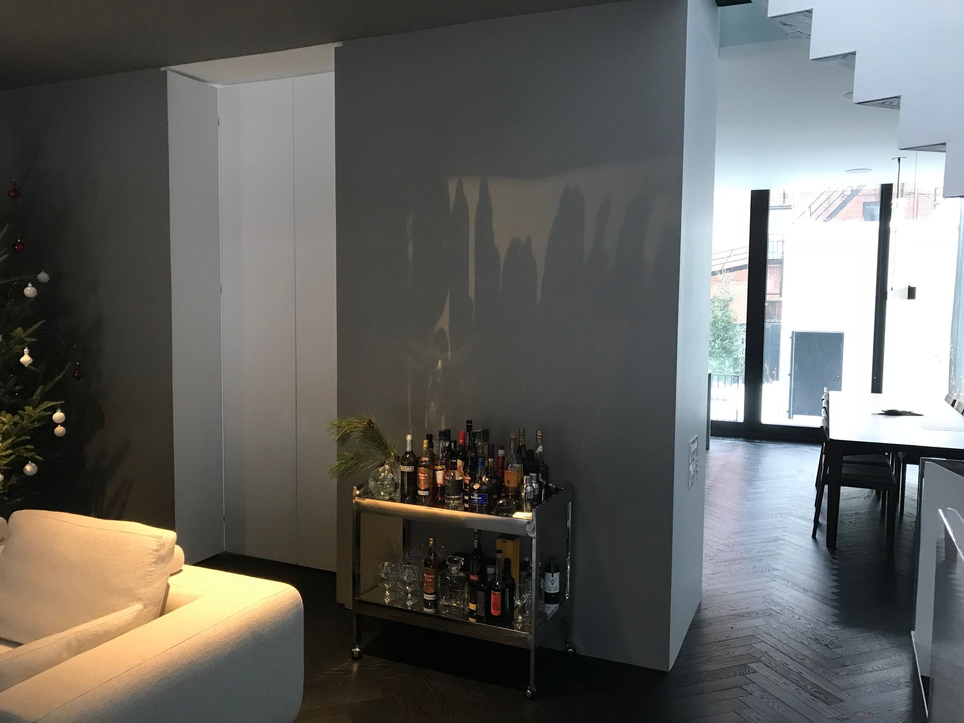 Intérieur moderne avec un canapé beige, un chariot de bar avec bouteilles et une vue dégagée sur un couloir lumineux.