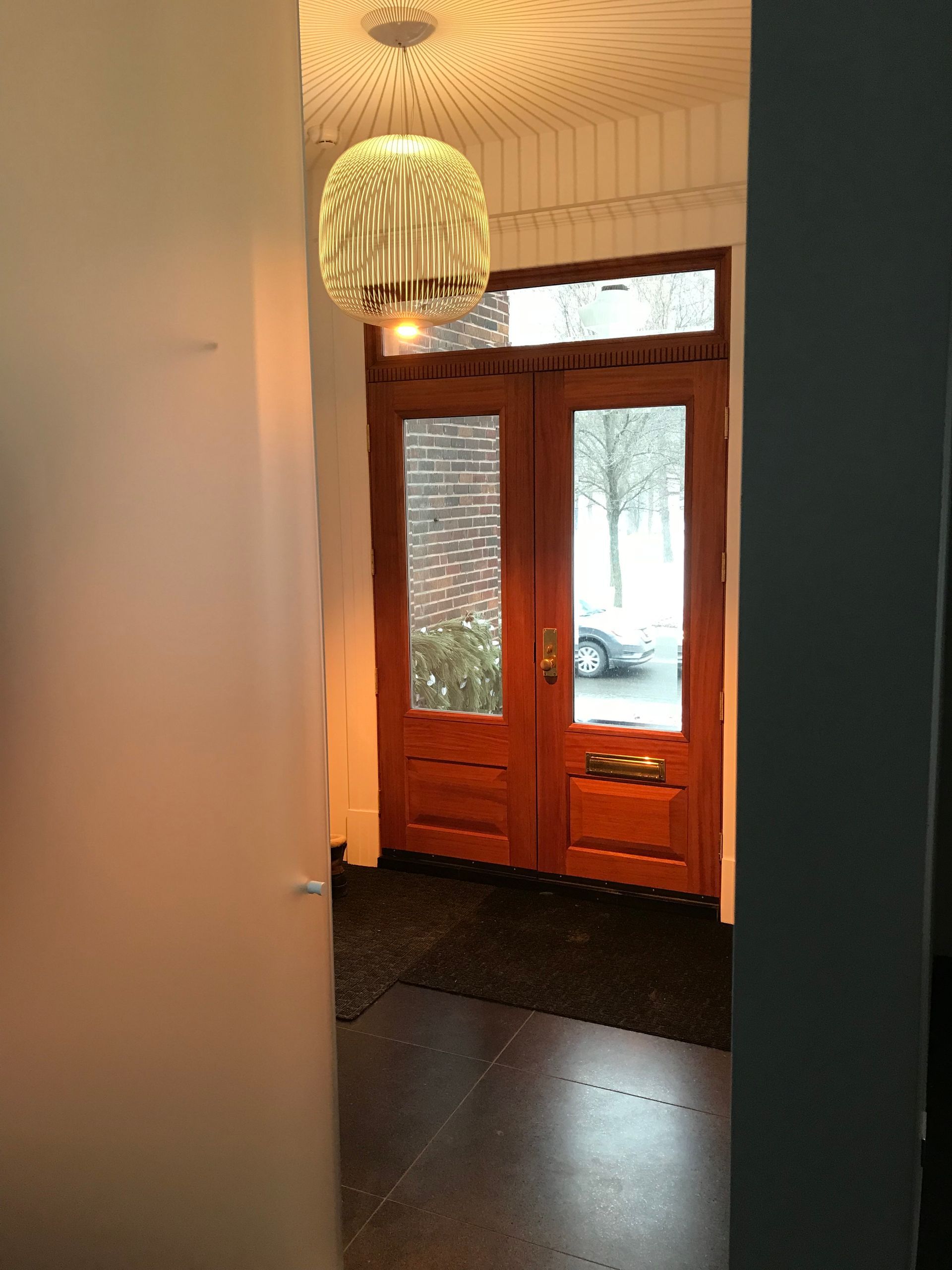 Couloir avec une porte d'entrée en bois, des panneaux de verre à motifs et un luminaire suspendu.