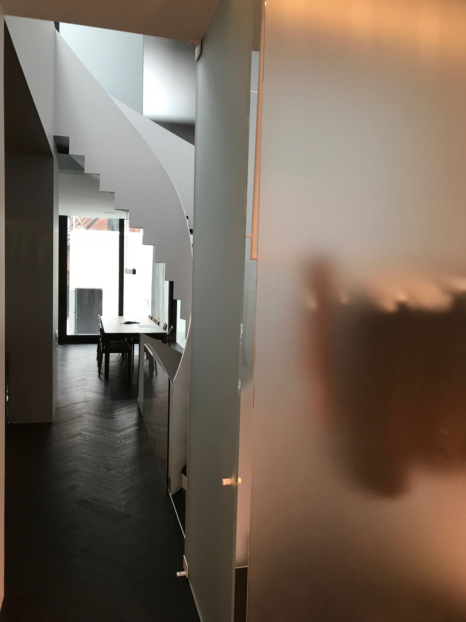 Couloir étroit avec un escalier blanc incurvé, une paroi en verre dépoli et une petite table près d'une fenêtre lumineuse.