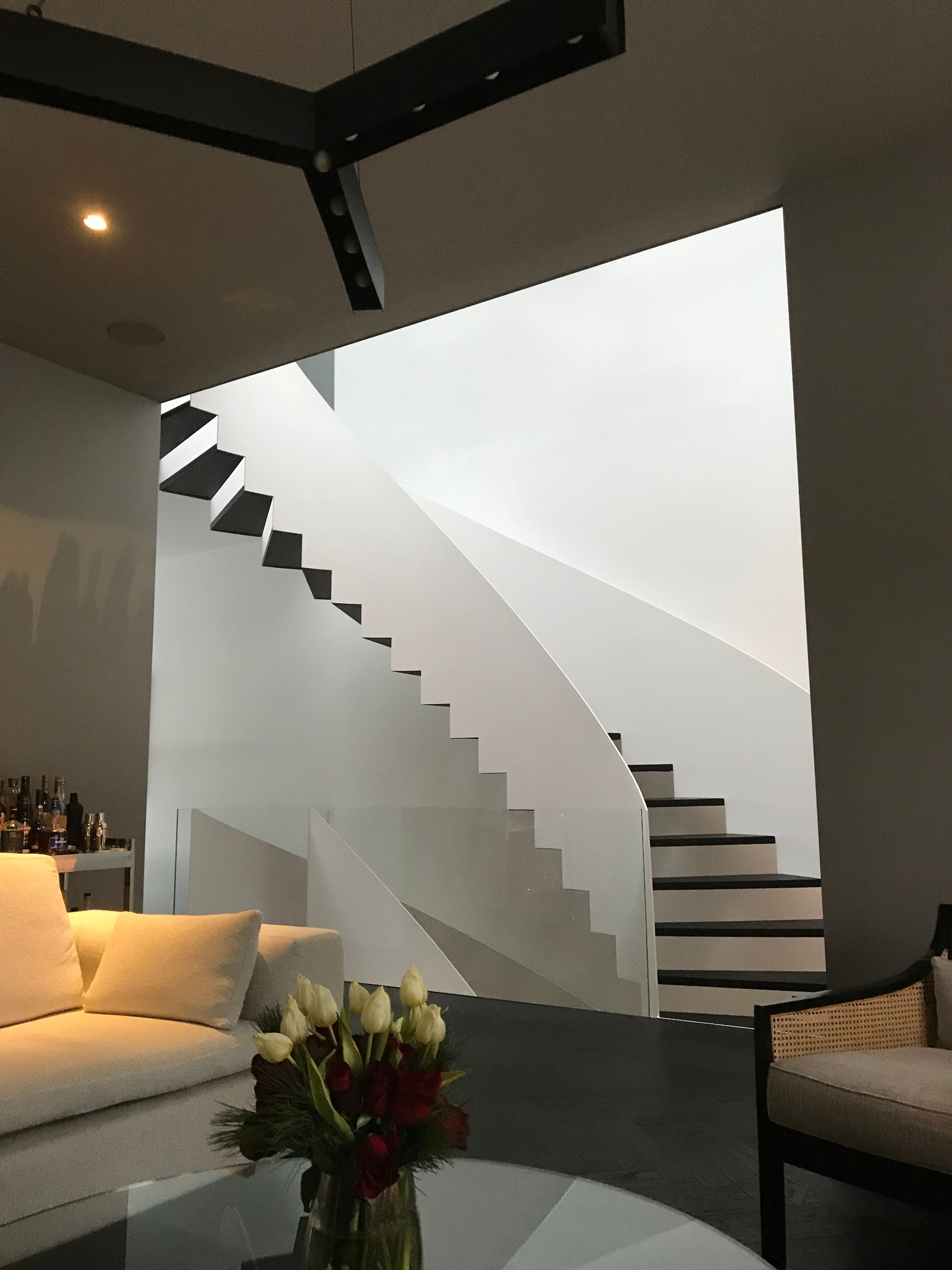 Salon moderne avec escalier blanc incurvé, murs gris, canapé et table basse ornée de fleurs