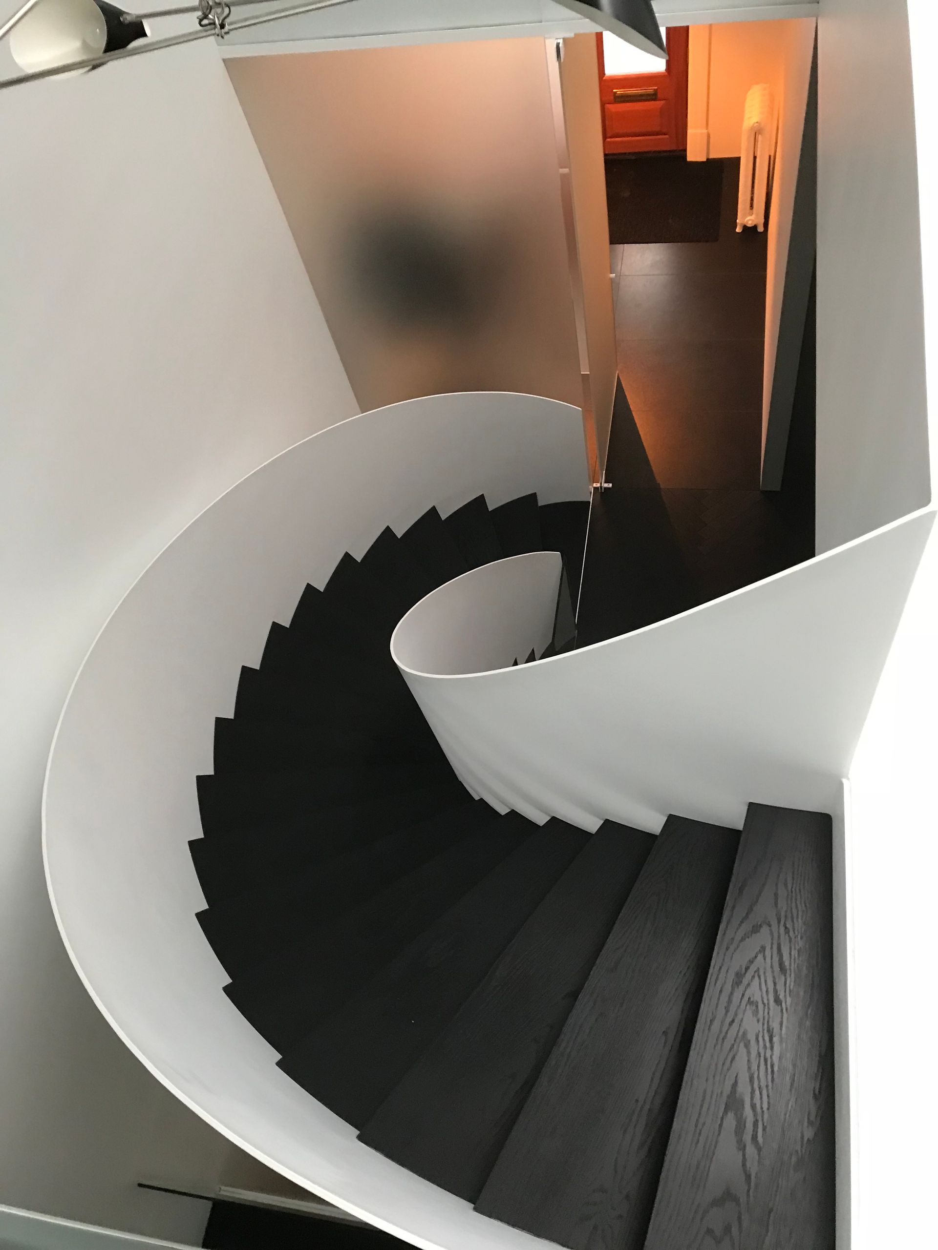 Escalier en colimaçon aux marches noires et aux murs blancs, dans un intérieur moderne, menant à une porte baignée d'une lumière chaude.