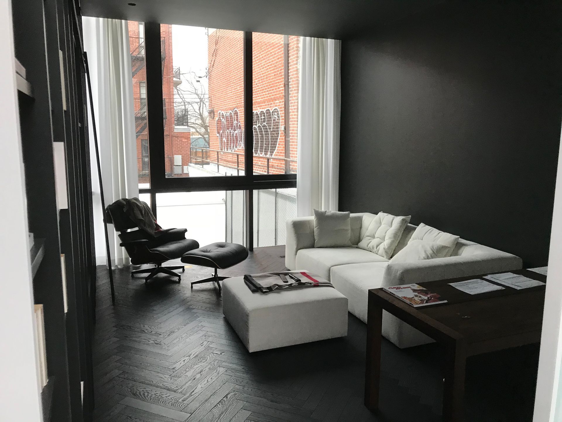 Salon moderne avec canapé blanc, fauteuil noir, table basse et grande fenêtre offrant une vue sur la ville.