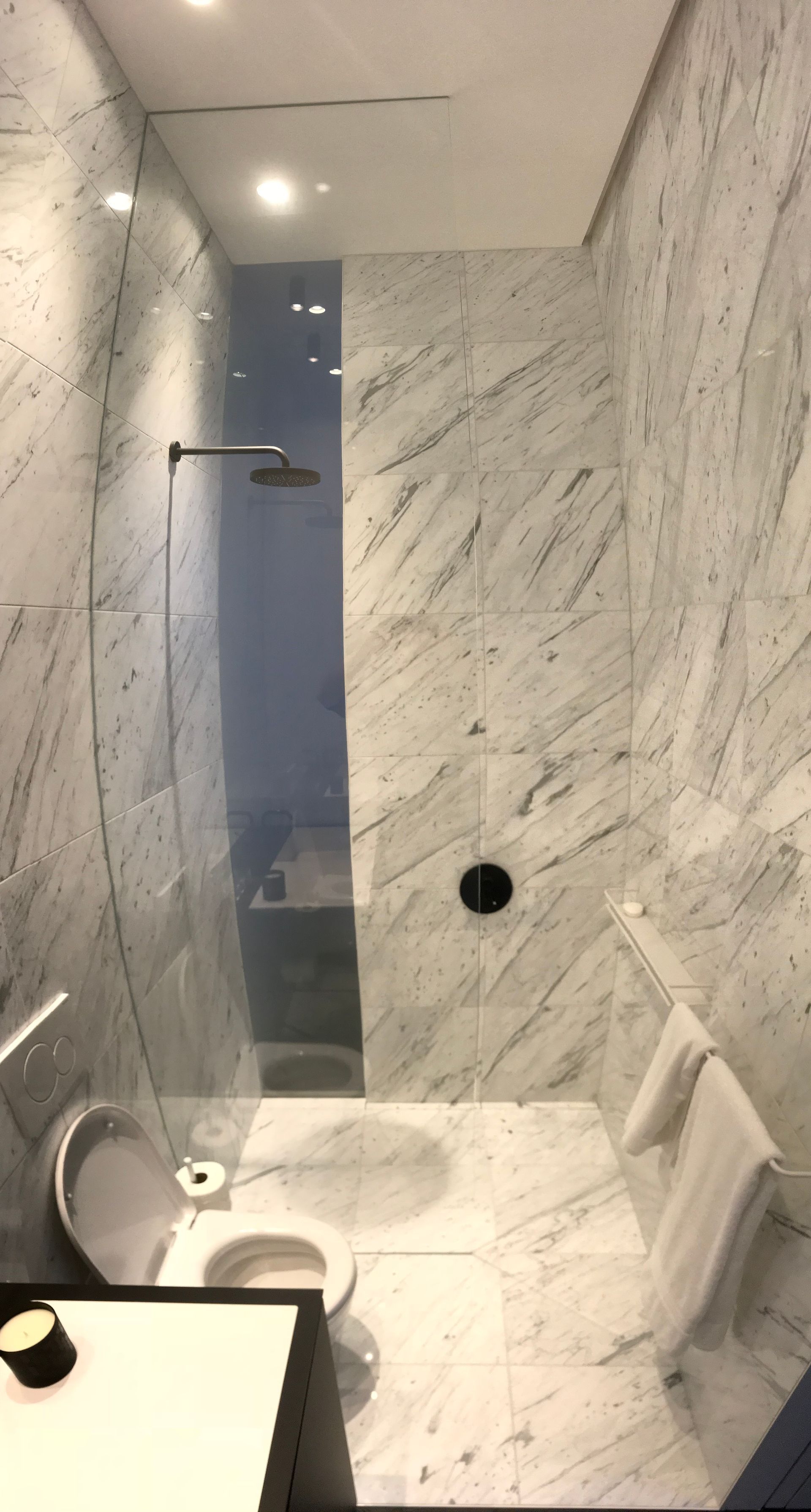 Salle de bains moderne carrelée en marbre avec douche en verre, pommeau de douche mural, toilettes et lavabo dans un espace étroit