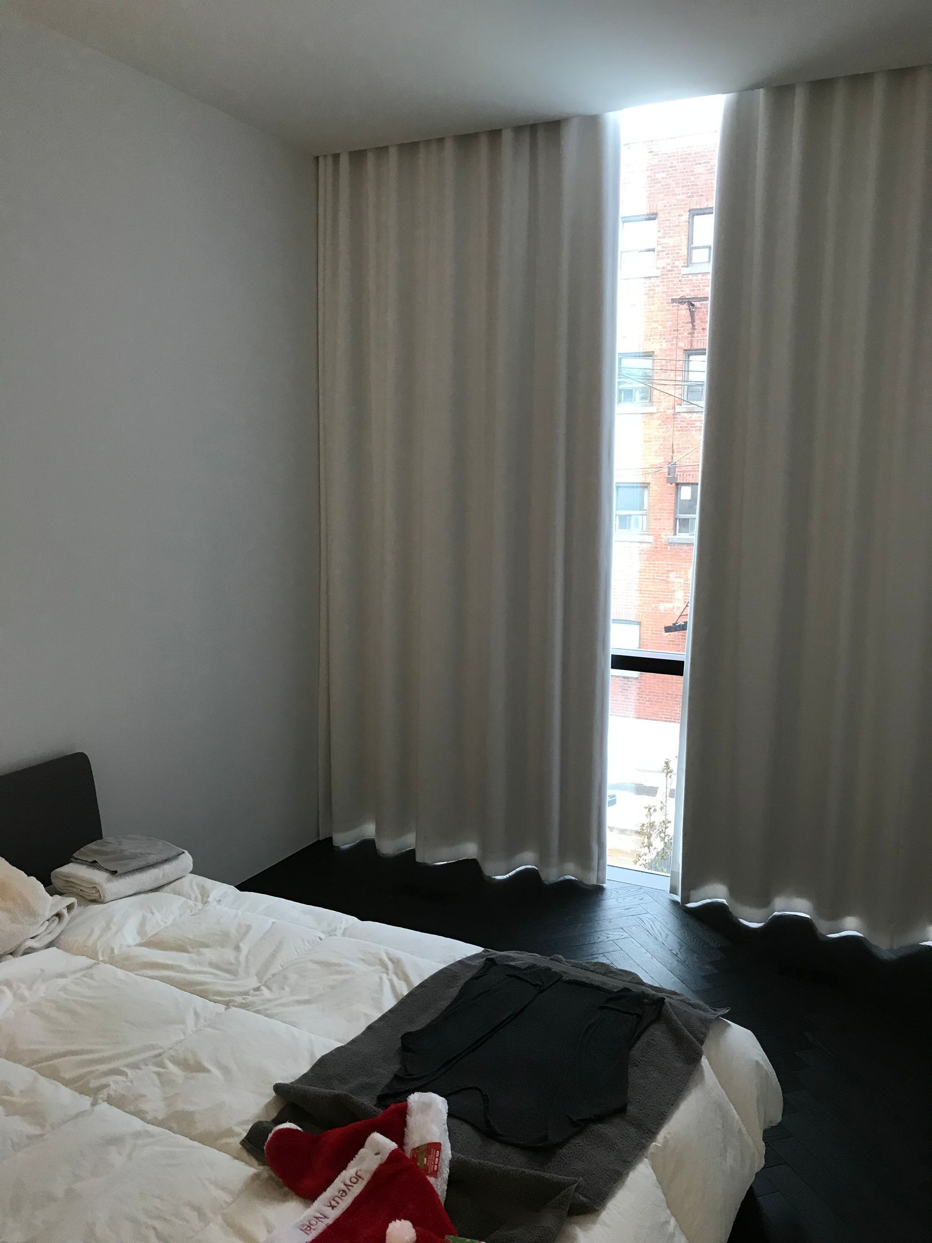 Chambre avec lit défait, rideaux sombres et vue sur des bâtiments en briques depuis une fenêtre étroite