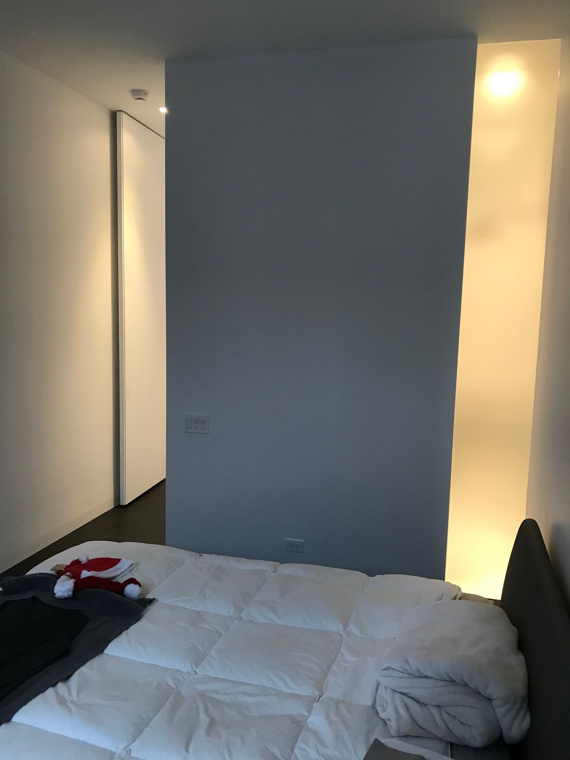 Chambre minimaliste avec un lit défait, un mur d'accent bleu et des lumières chaudes dans le couloir.