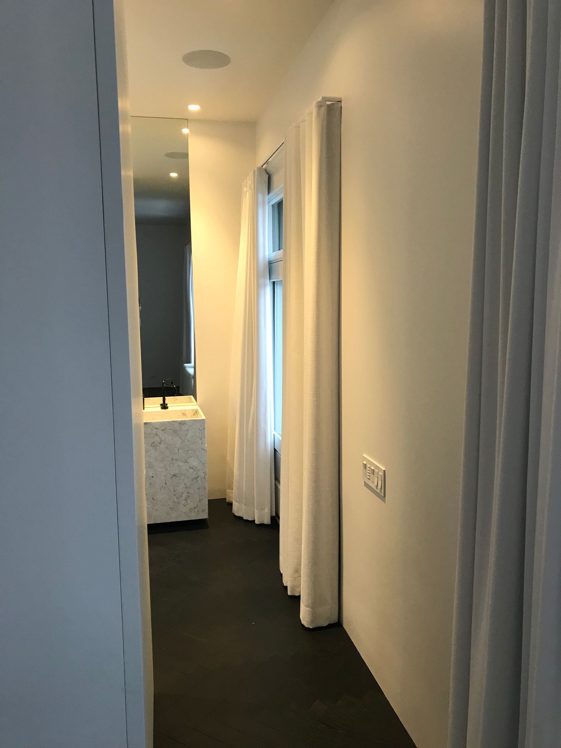 Couloir étroit aux murs blancs, moquette foncée, rideaux et une porte au fond.
