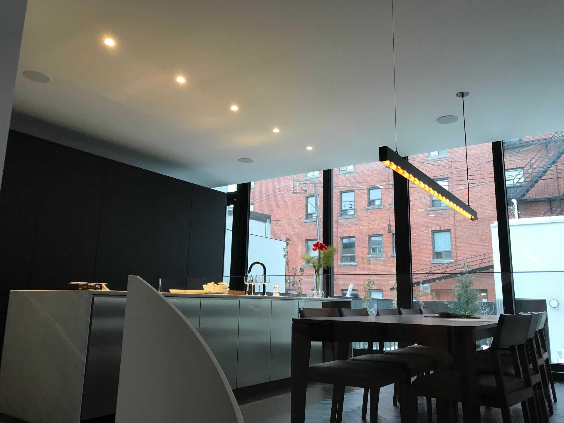 Cuisine et salle à manger modernes avec placards noirs, îlot central blanc et grandes fenêtres donnant sur un bâtiment en briques