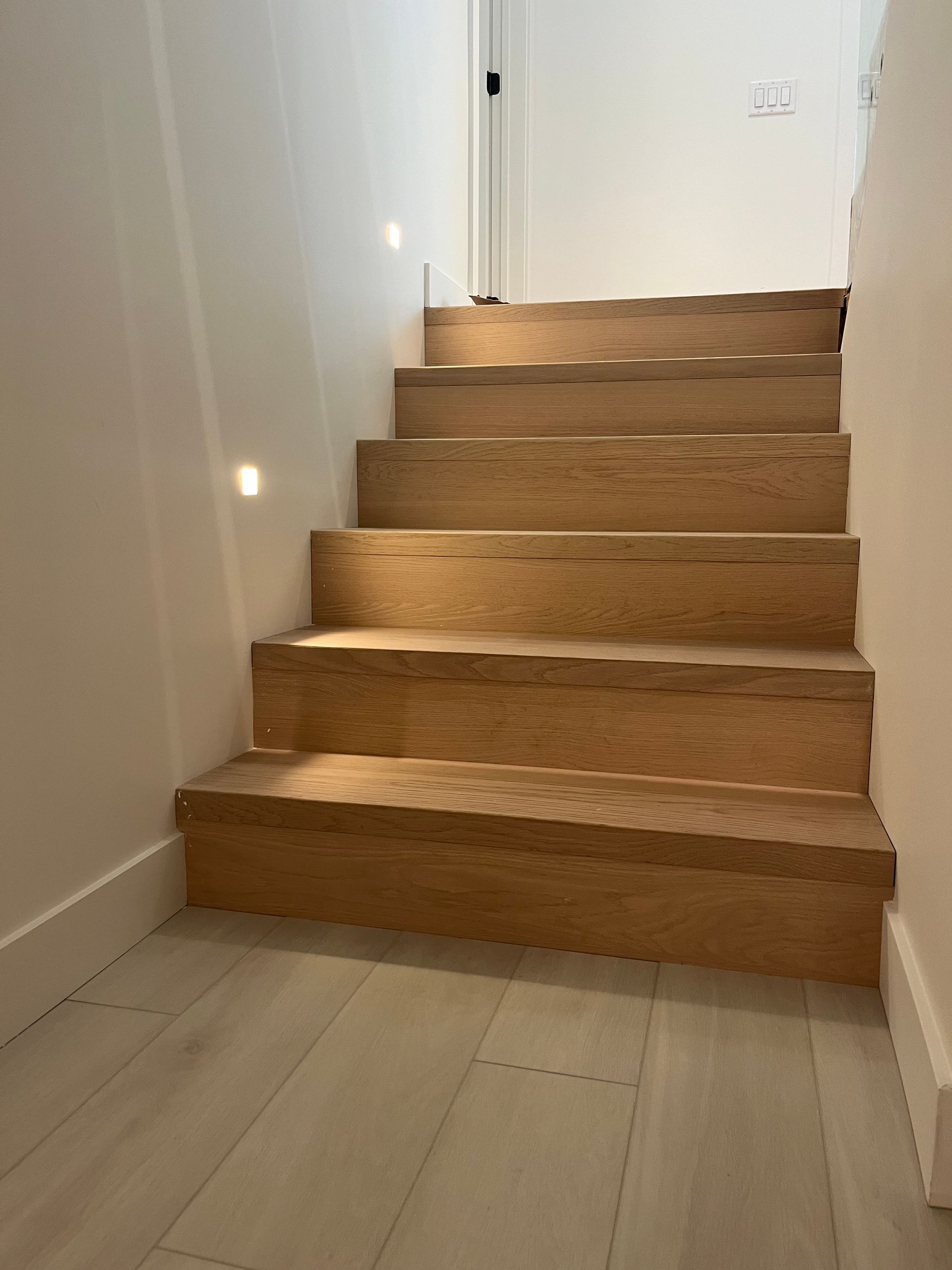 Un escalier intérieur étroit aux marches en bois et aux murs blancs mène à une porte fermée.