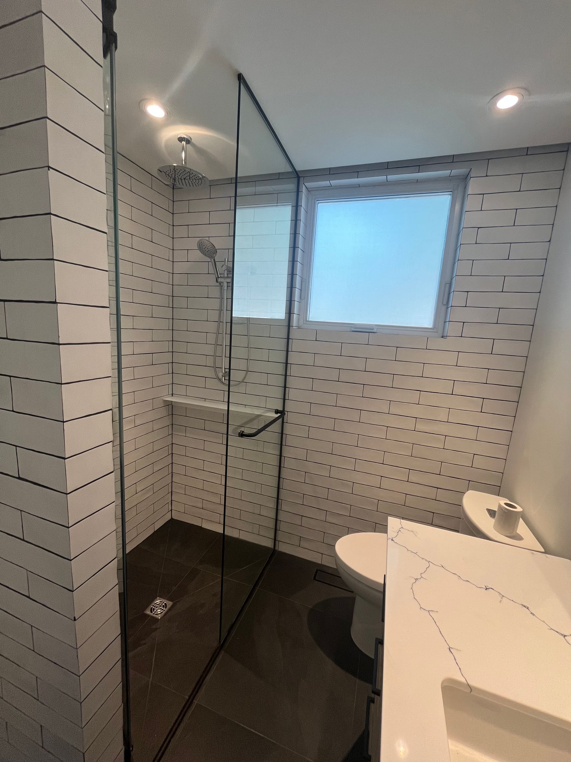 Salle de bain moderne carrelée avec douche en verre, fenêtre et lavabo blanc