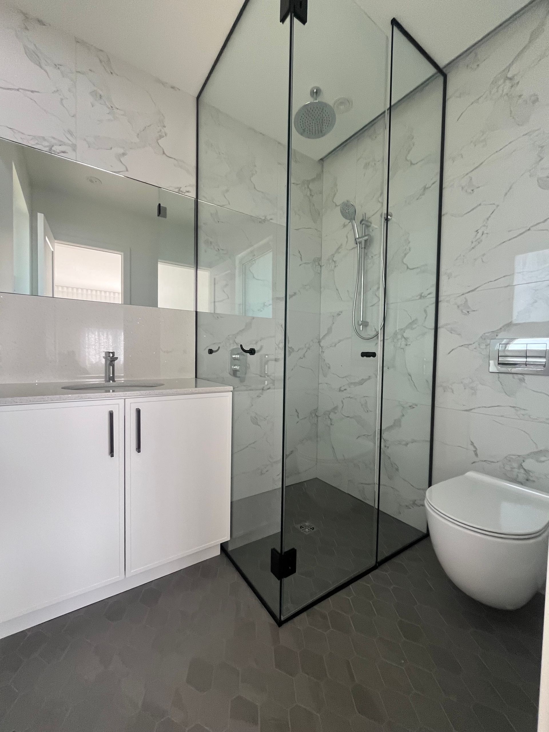 Salle de bains moderne avec douche en verre, meuble-lavabo blanc et toilettes contre des murs à motif marbre.
