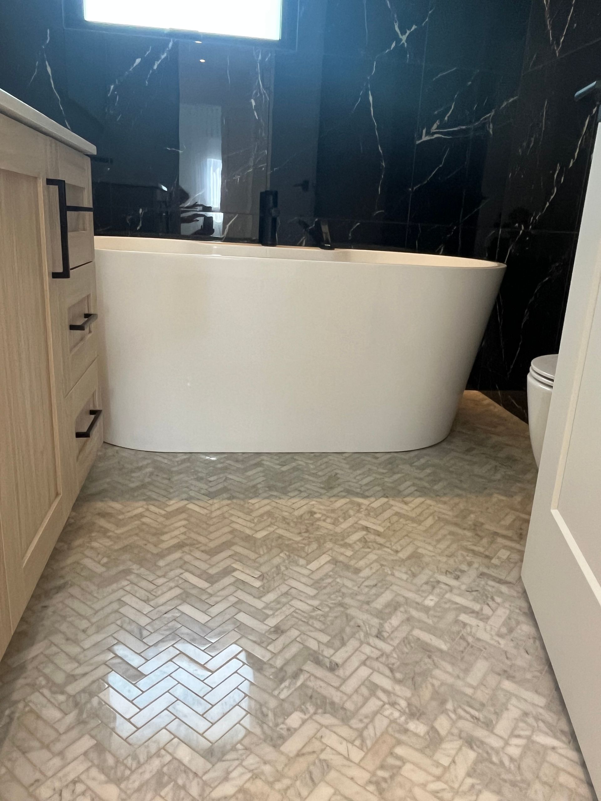 Salle de bains moderne avec baignoire blanche autoportante, mur en marbre noir et sol en carrelage à chevrons.