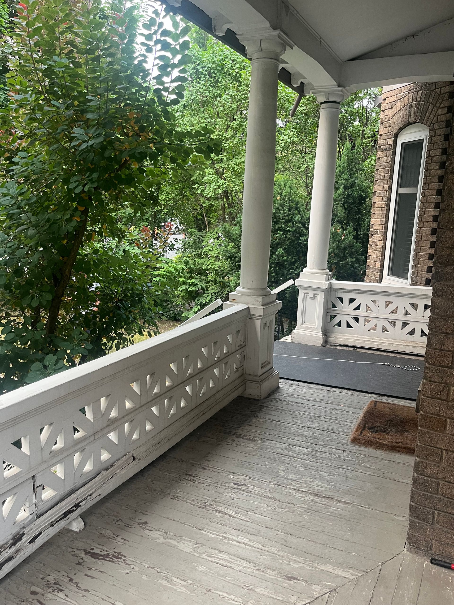 Porche couvert avec balustrades et colonnes blanches, bordé d'arbres feuillus et d'une maison en briques