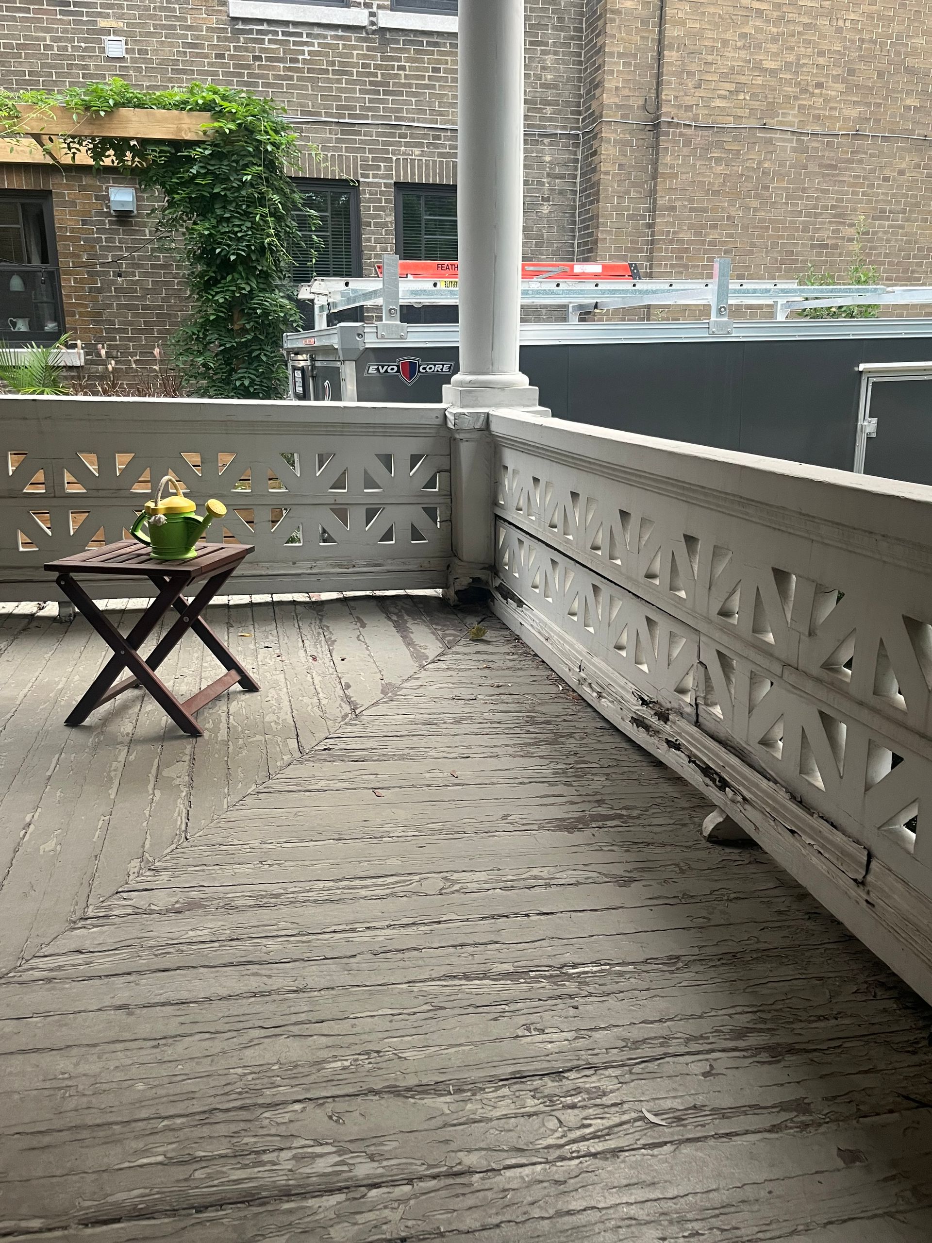 Petit patio avec un support à plantes pliable, une balustrade blanche et un mur en pierre en arrière-plan.