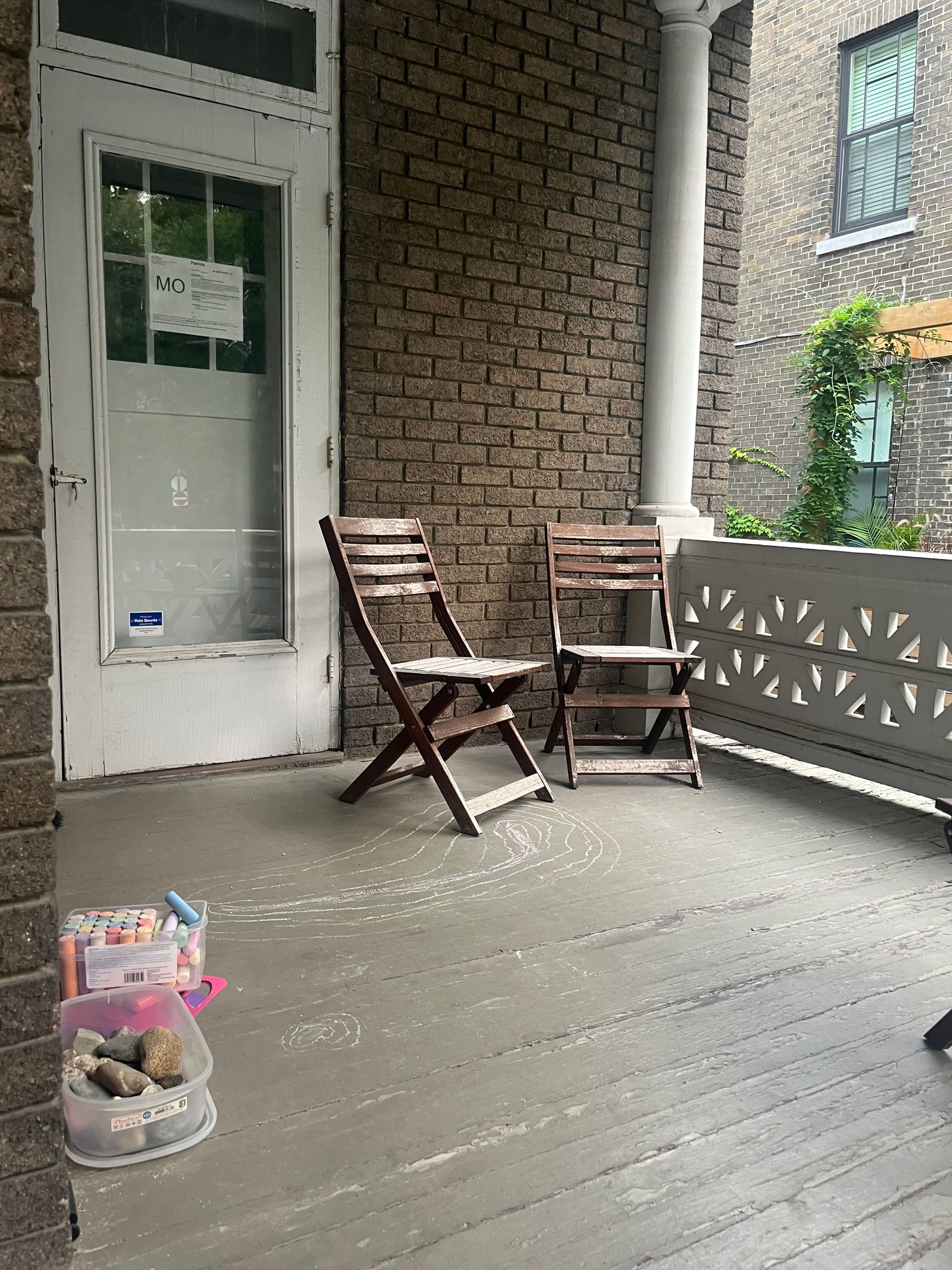 Porche d'entrée avec deux chaises en bois, une porte blanche et une jardinière contre un mur de briques.