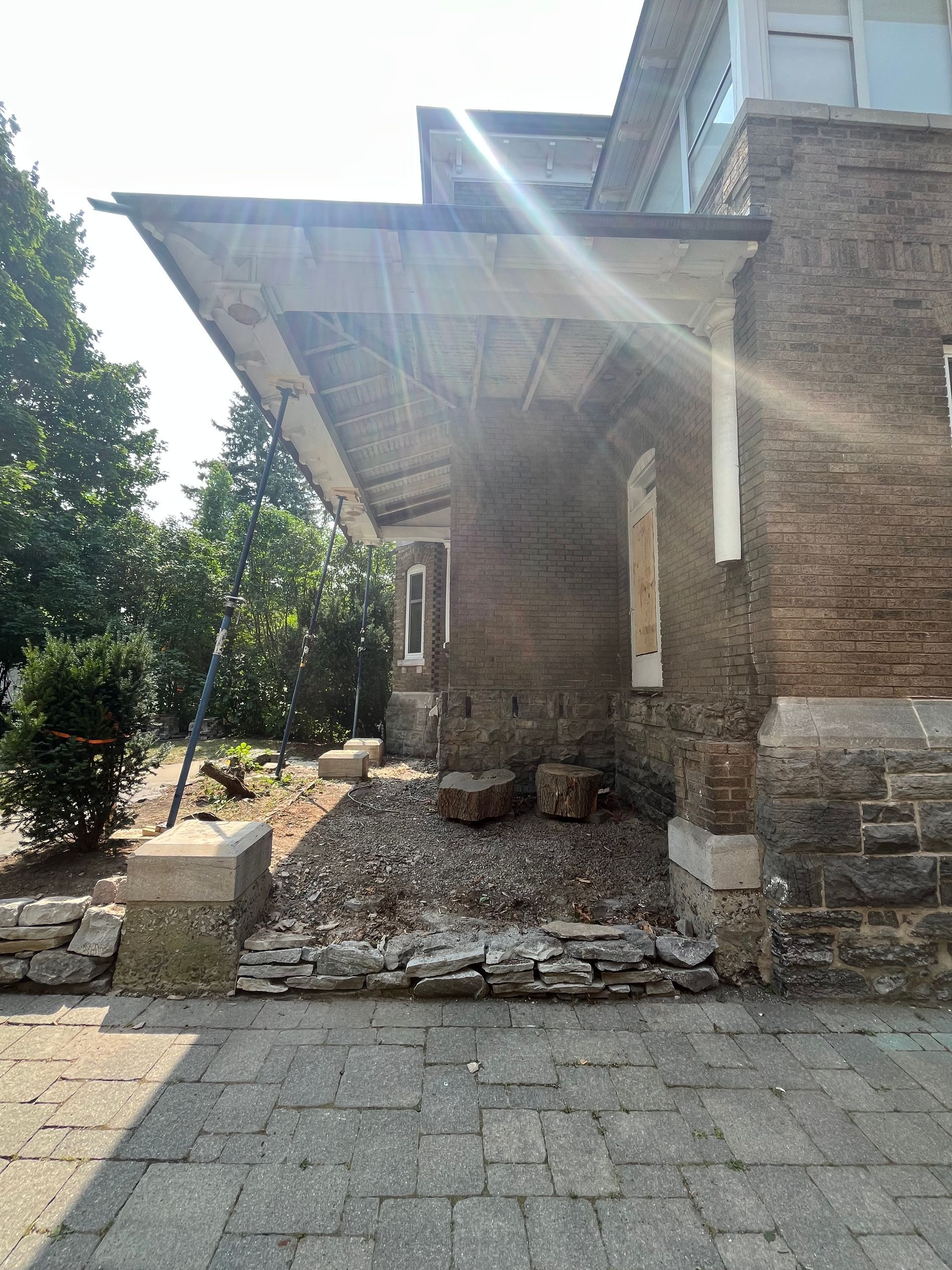 Entrée d'un bâtiment en pierre avec un auvent métallique, un mur de briques et une allée pavée ensoleillée devant.