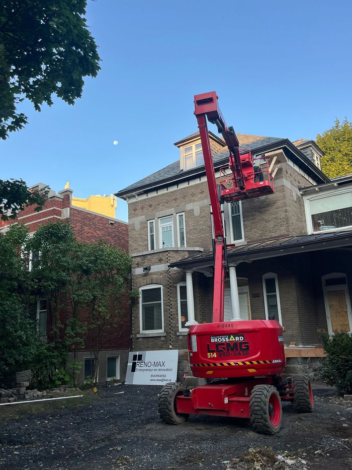 Nacelle élévatrice rouge stationnée près d'une maison en rénovation, avec des ouvriers sur la partie supérieure du toit.