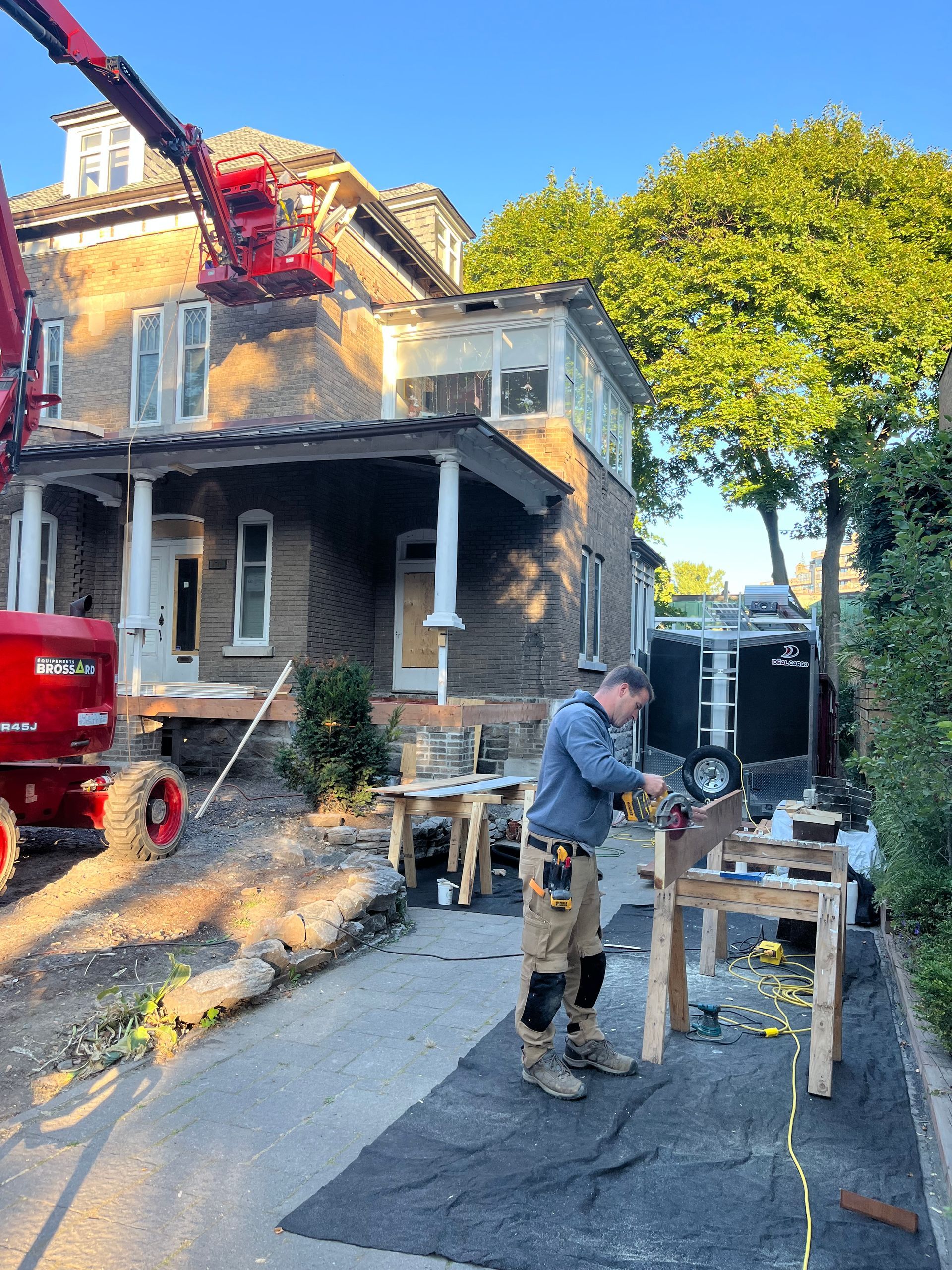 Des ouvriers du bâtiment travaillent à l'extérieur d'une maison, avec une nacelle élévatrice rouge et des débris dans l'allée.