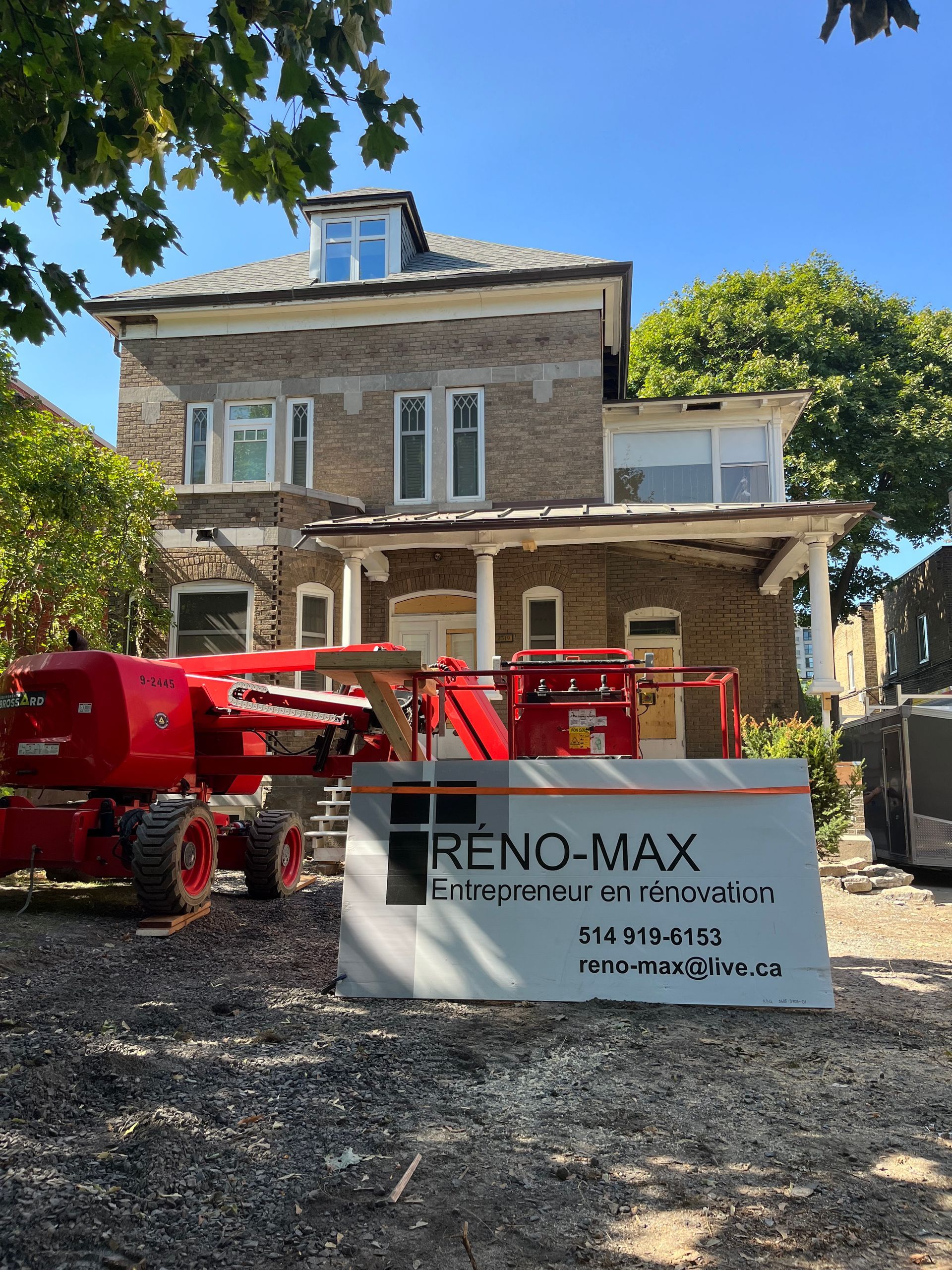 Maison en pierre avec nacelle élévatrice rouge et panneau RENO-MAX dans la cour avant sous un ciel bleu azur