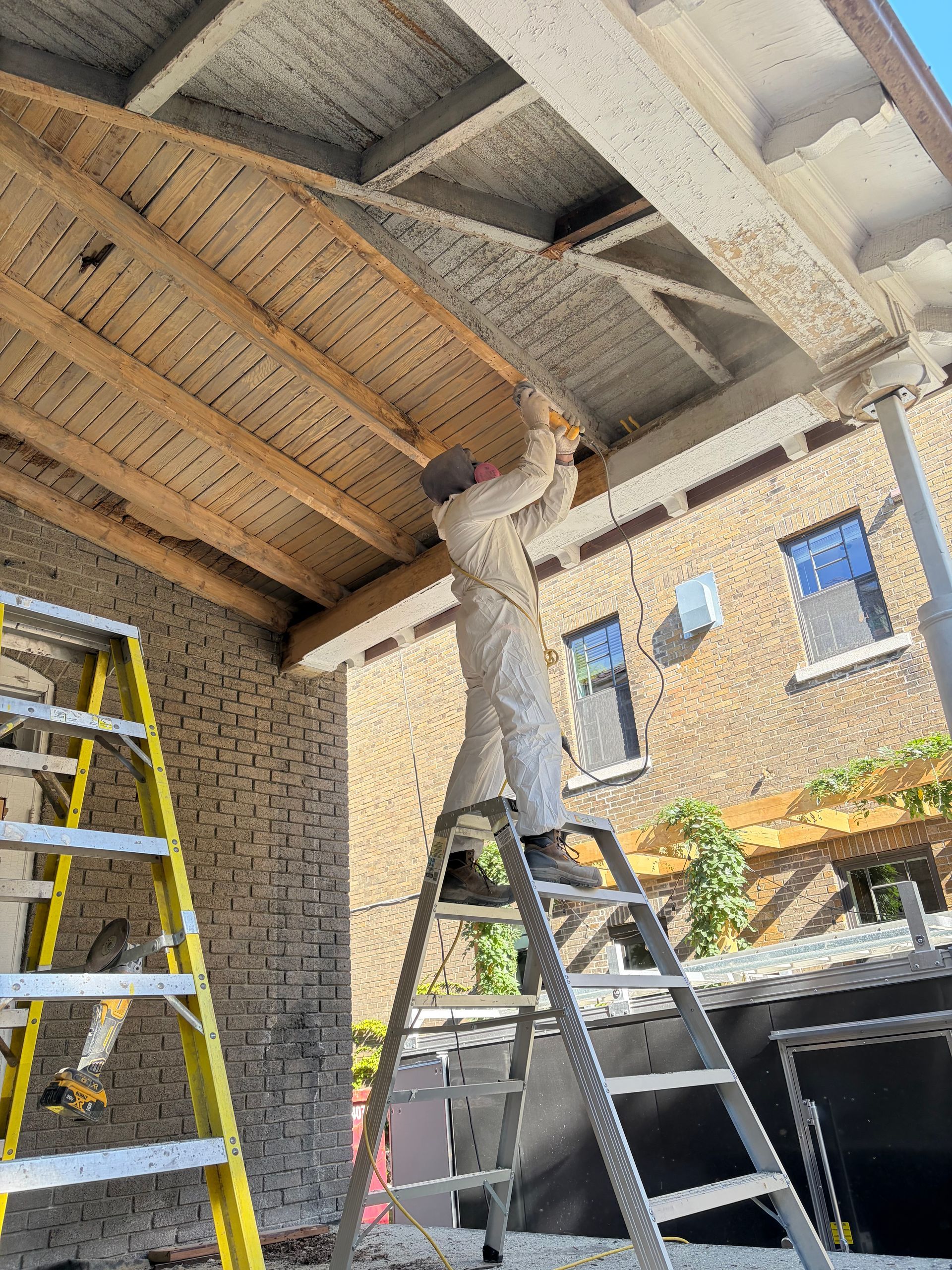 Une personne perchée sur une échelle peint un mur de briques sous un patio couvert dont les poutres du plafond sont apparentes.