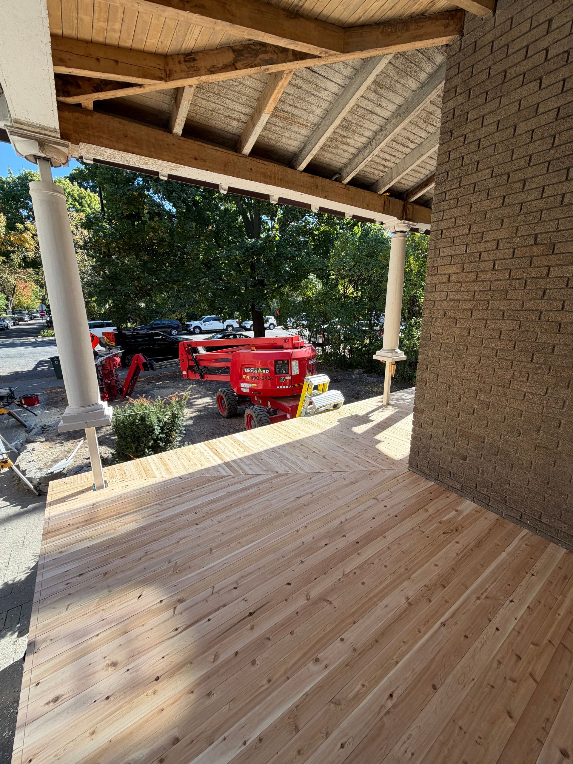 Patio couvert avec plafond en bois, colonnes blanches et une nacelle élévatrice rouge à l'extérieur, au milieu des arbres.