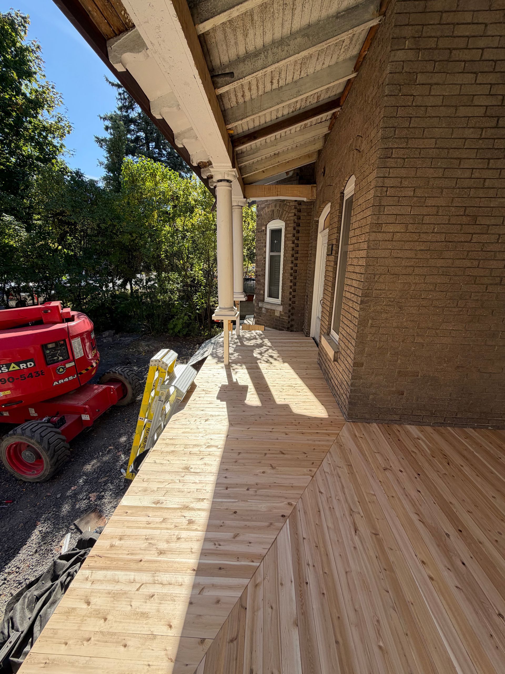 Porche en bois en construction à côté d'une maison en briques, avec un ascenseur rouge et une allée en gravier.