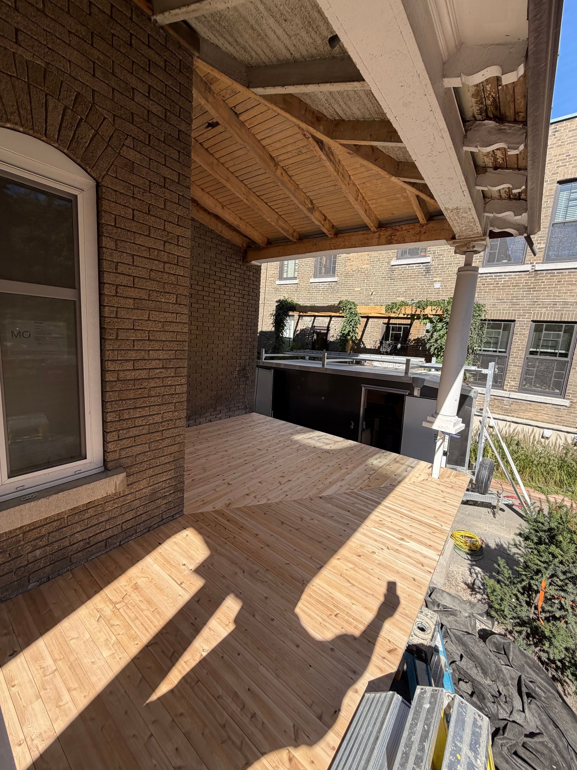 Porche couvert avec mur de briques, fenêtre, plancher en bois et ombres baignées de soleil.