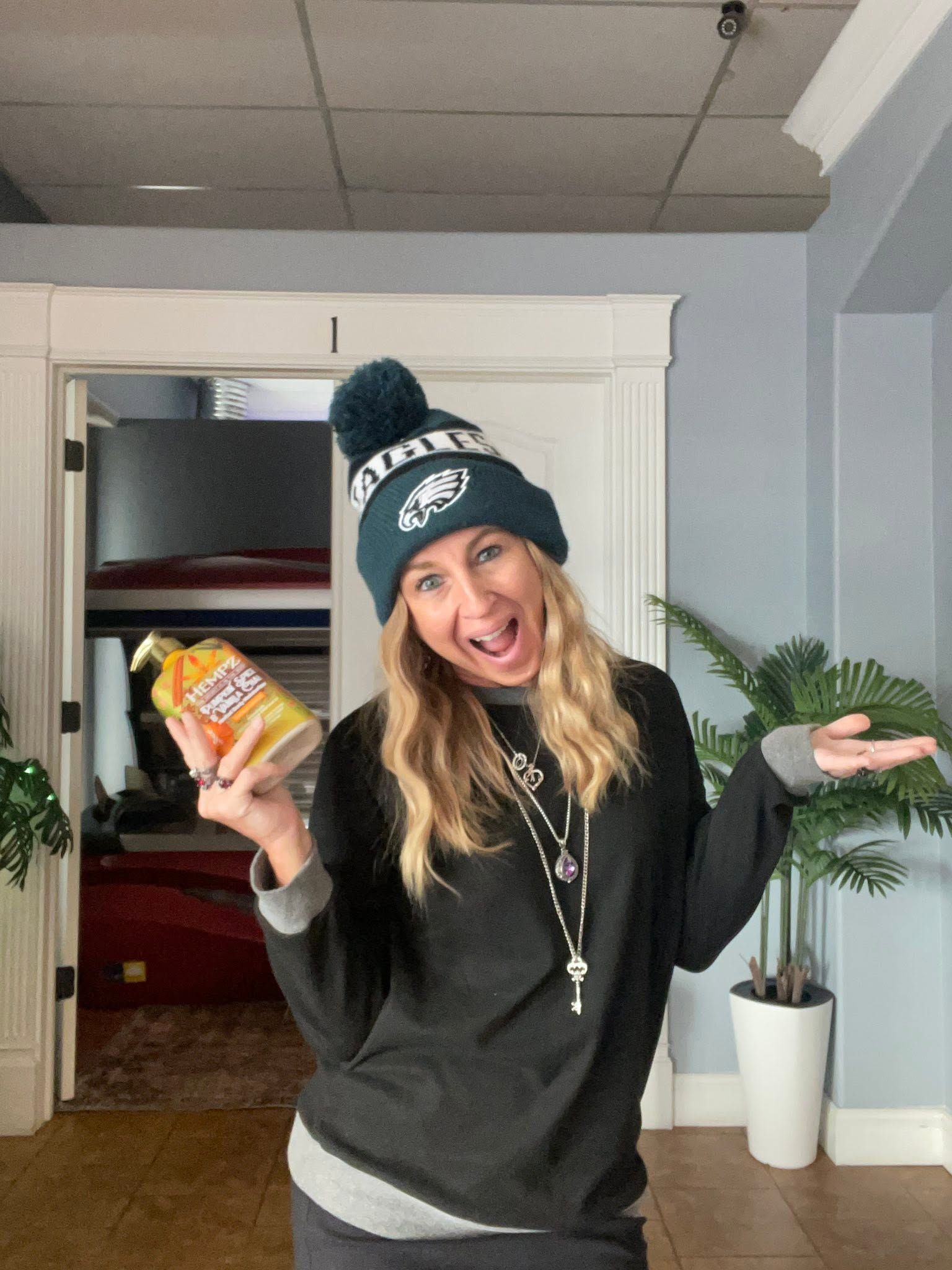 Woman in black sweater and teal beanie smiling indoors, holding a snack bag and gesturing beside a plant