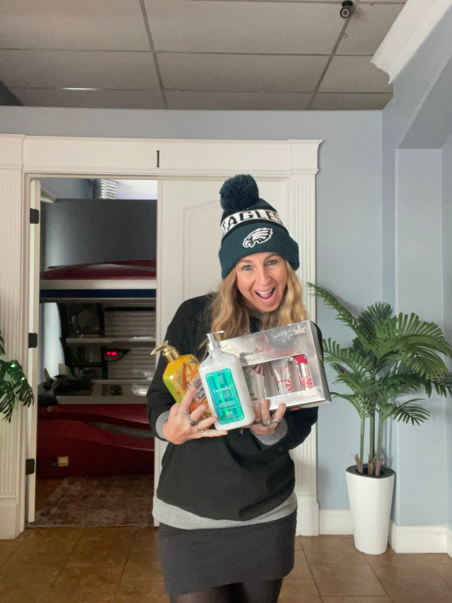 Woman holding boxed items in a doorway, smiling in a gray room with a potted plant.