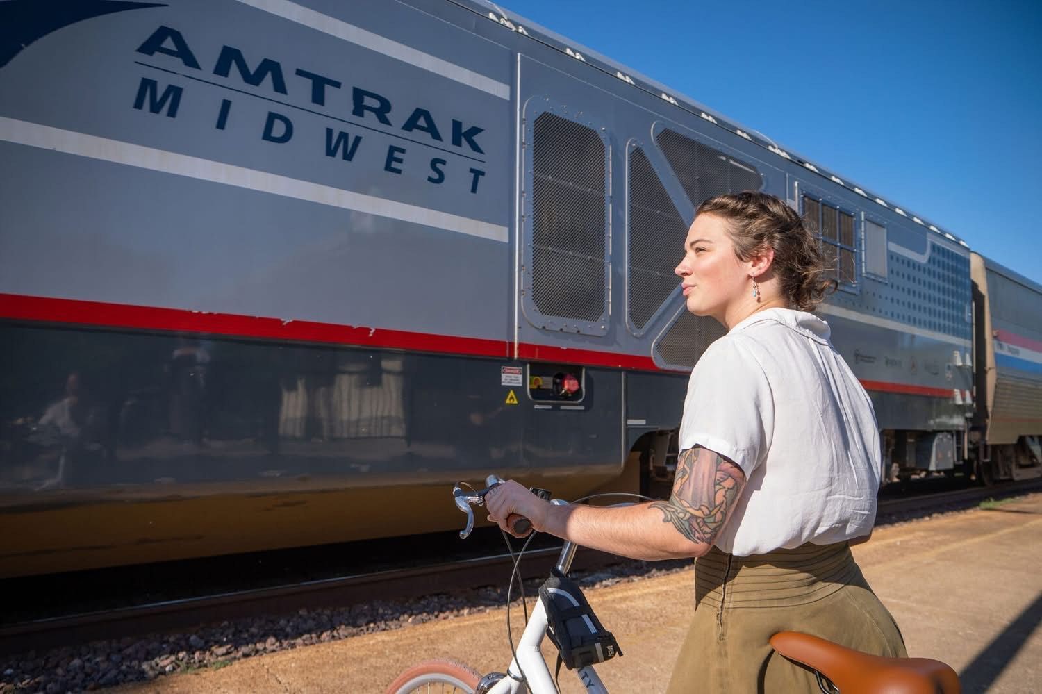 Woman with bicycle near Amtrak Midwest train on sunny day.