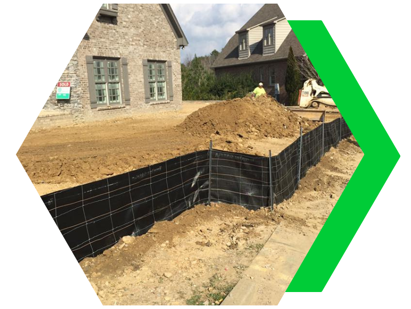 Construction site with black silt fencing and dirt near stone houses, marked by a green arrow.