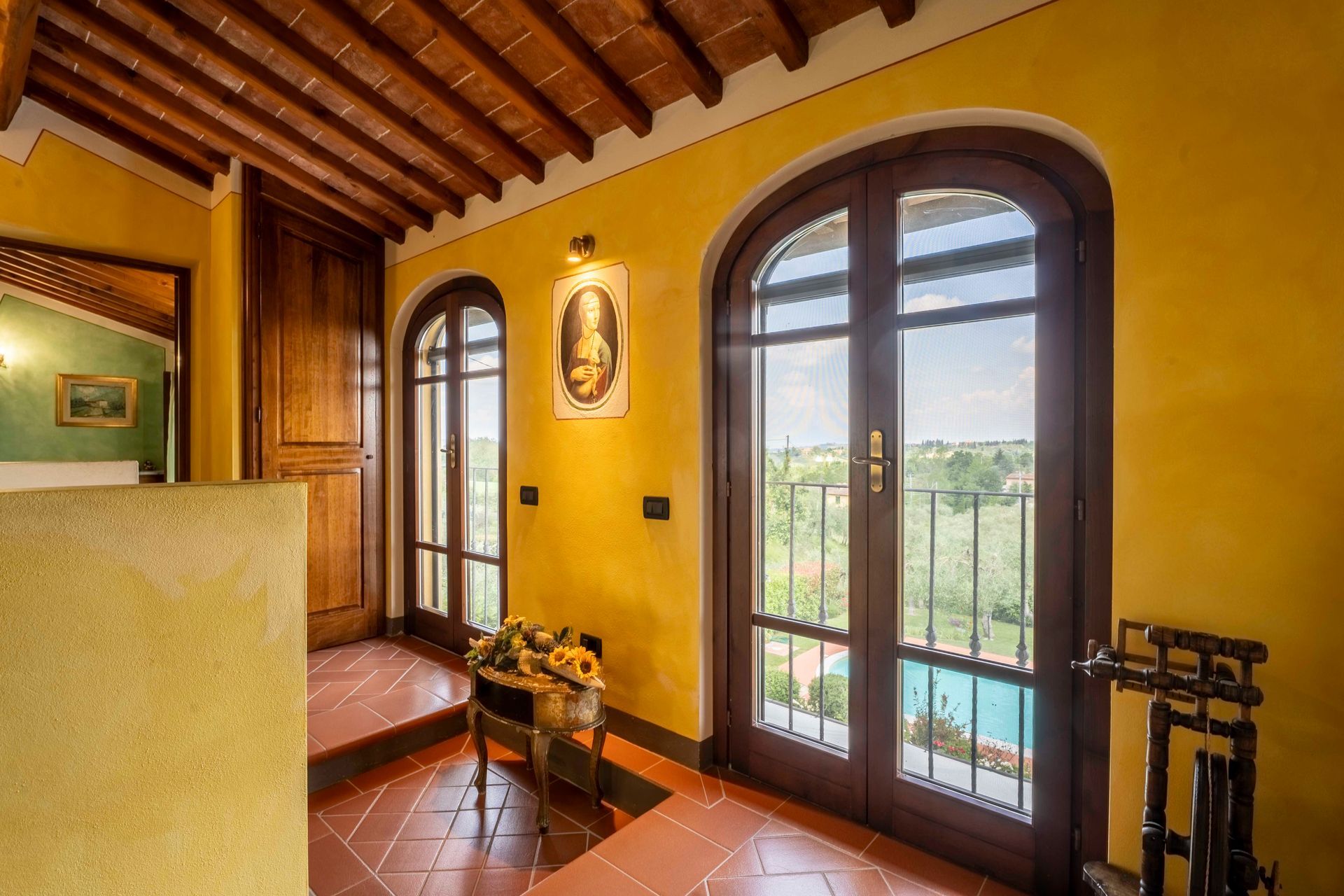 Accogliente corridoio con finestre ad arco, pavimento in cotto, travi in legno e vista dal balcone oltre le porte a vetri.