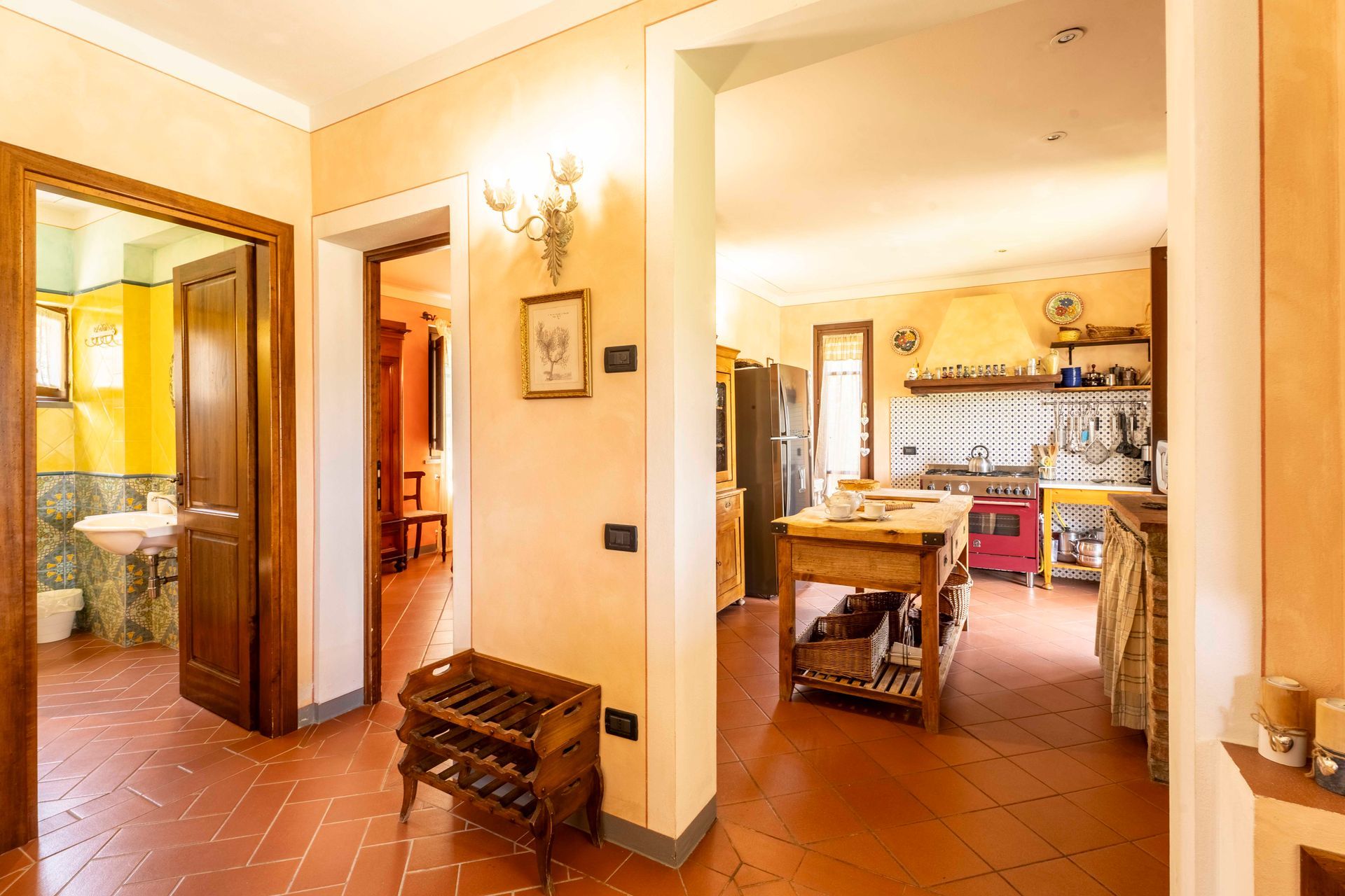 Corridoio e cucina dai toni caldi con pavimenti in piastrelle di terracotta, mobili in legno e porte aperte.