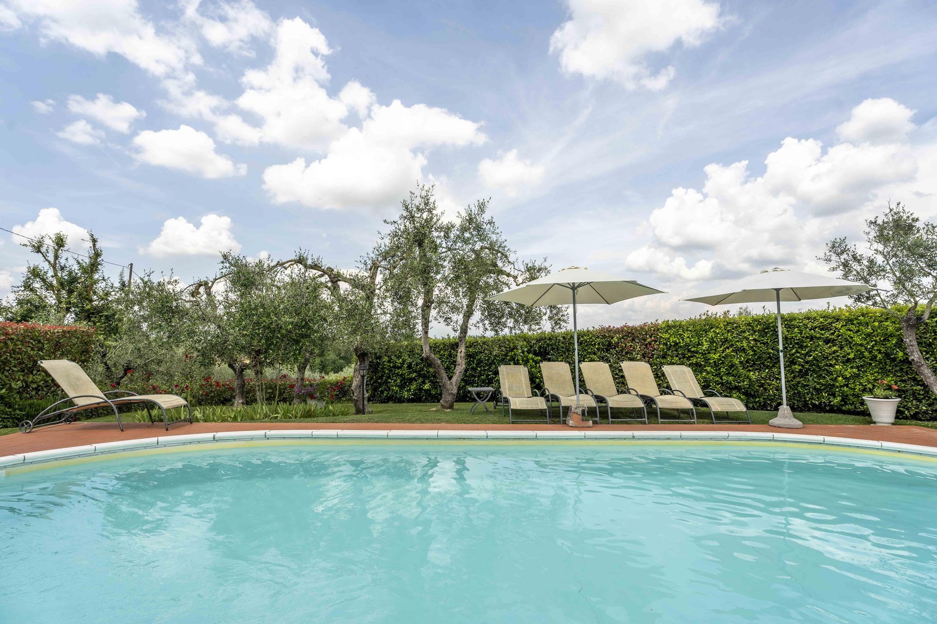 Piscina all'aperto con lettini, ombrelloni bianchi e vegetazione curata sotto un cielo nuvoloso.