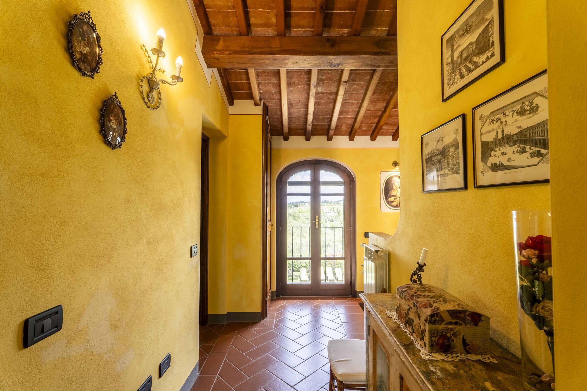 Stretto corridoio giallo con pavimento piastrellato, travi in legno a vista sul soffitto e porta ad arco in vetro in fondo.