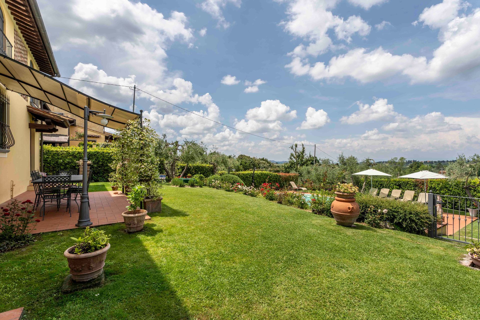 Un cortile soleggiato con prato verde, piante in vaso, posti a sedere in patio e un sentiero nel giardino sotto un cielo azzurro.