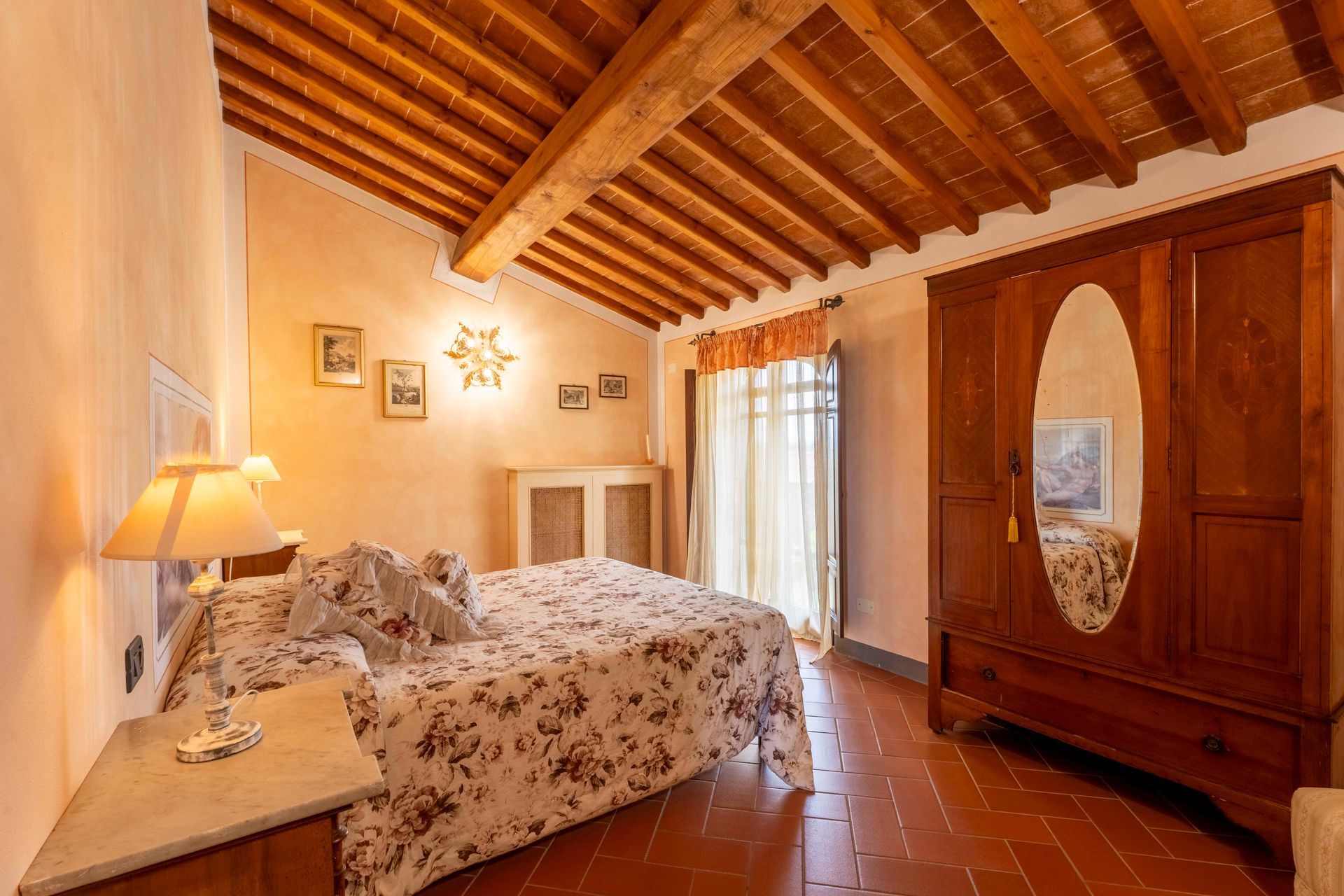 Accogliente camera da letto con copriletto floreale, travi a vista sul soffitto, armadio e tende trasparenti vicino alla finestra.