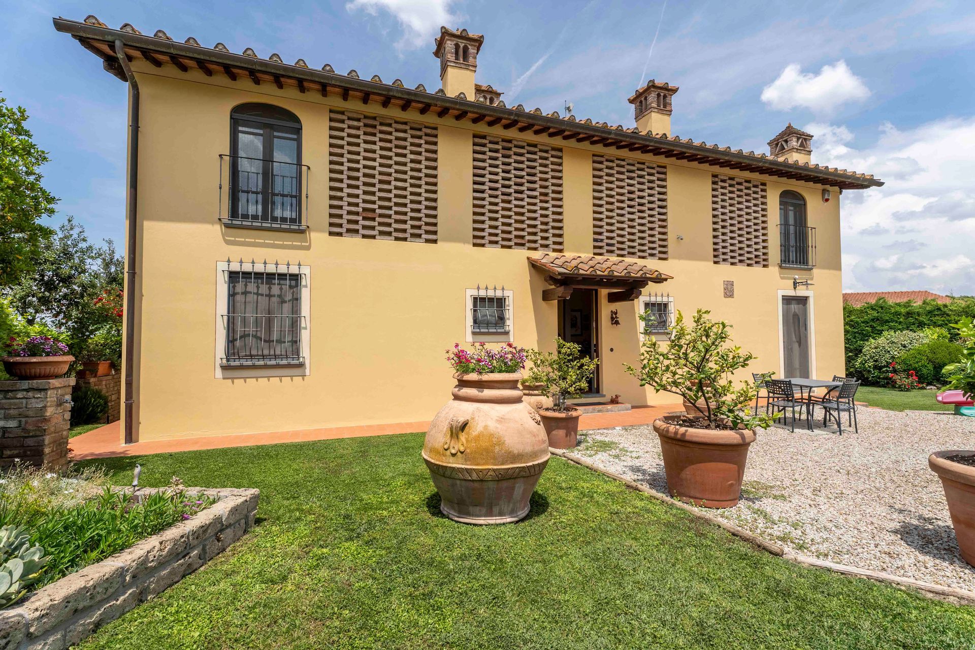 Casa gialla in stile mediterraneo con persiane, piante in vaso e un cortile di ghiaia sotto un cielo azzurro.