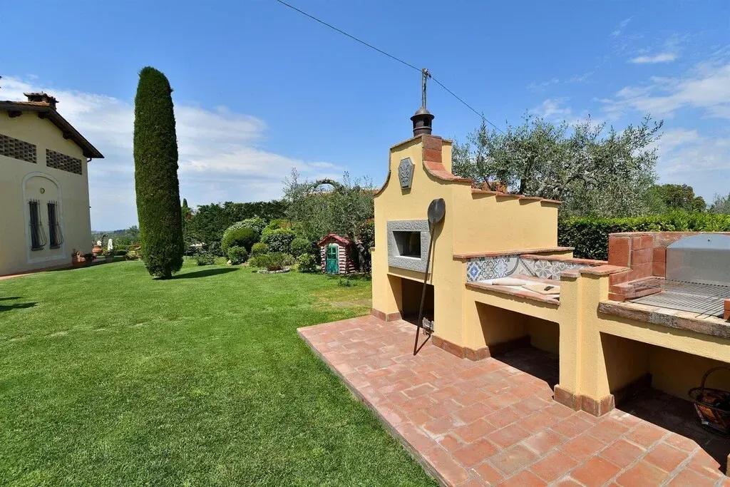 Cortile soleggiato con prato verde, patio piastrellato e barbecue in mattoni accanto a una casa in stucco giallo.