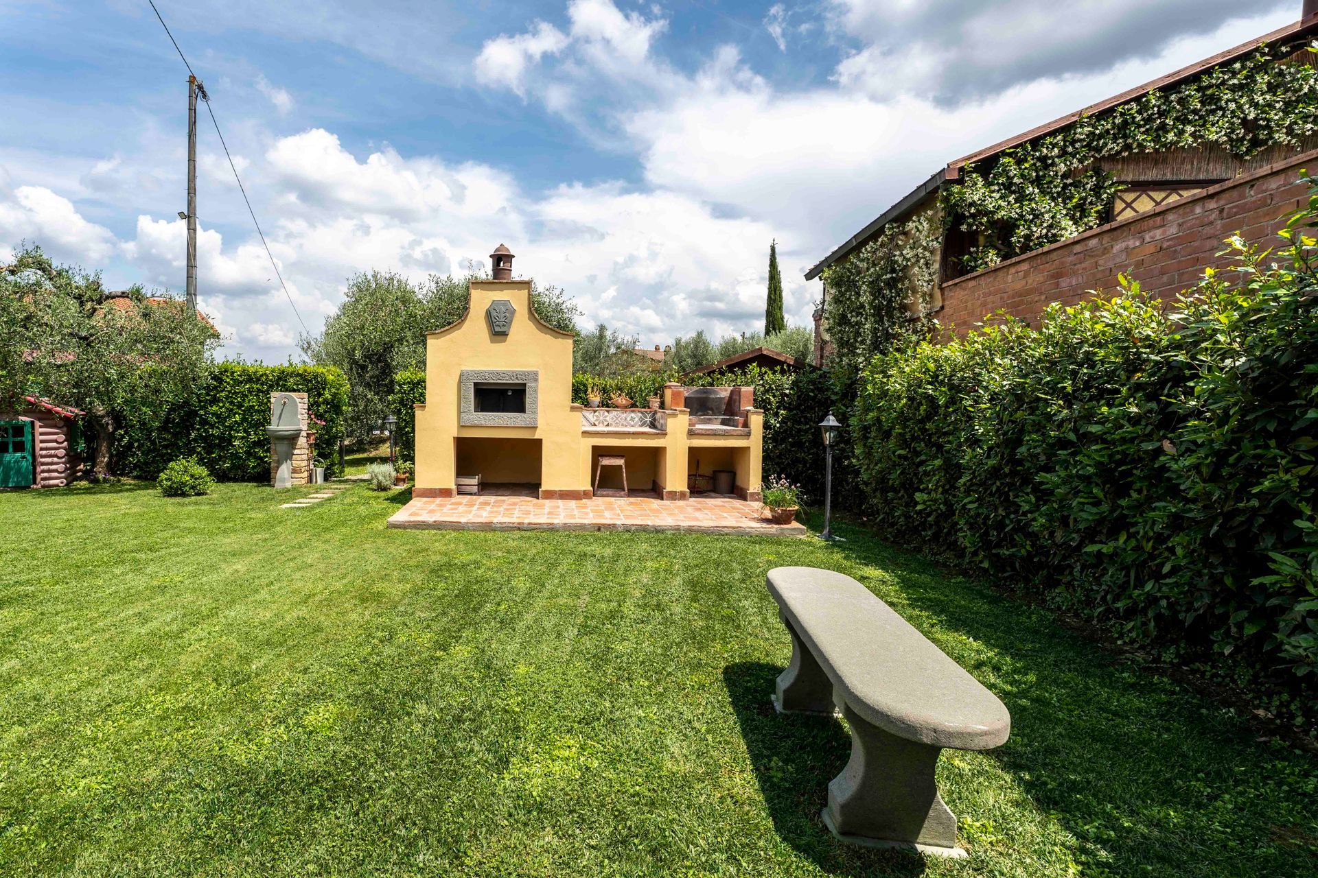 Un cortile soleggiato con un gazebo giallo, un prato verde, una panchina e piante rampicanti lungo un muro di mattoni.