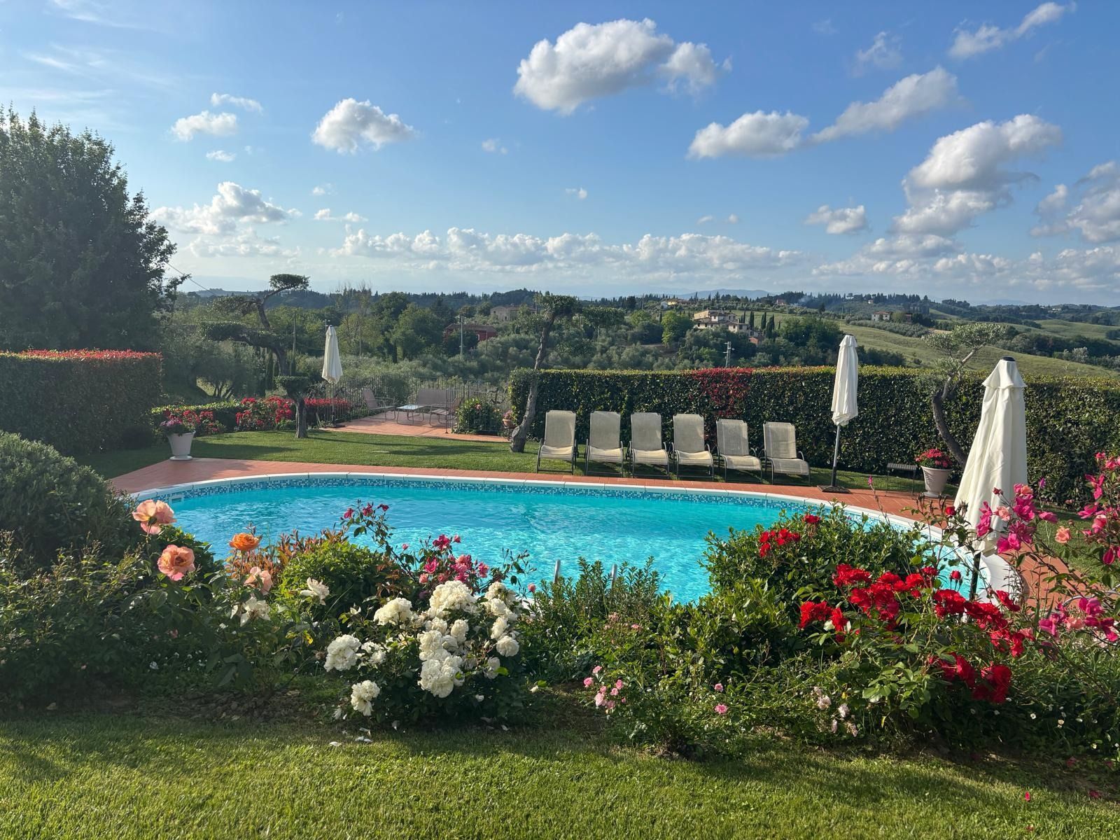 Piscina in giardino con acqua azzurra, fiori in fiore, lettini prendisole e una soleggiata vista sulla collina sotto un cielo parzialmente nuvoloso.
