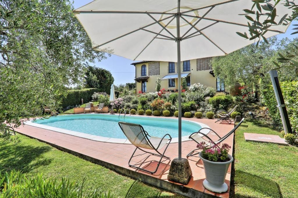 Piscina in giardino con ombrellone, lettini, vasi di fiori e una casa sullo sfondo.